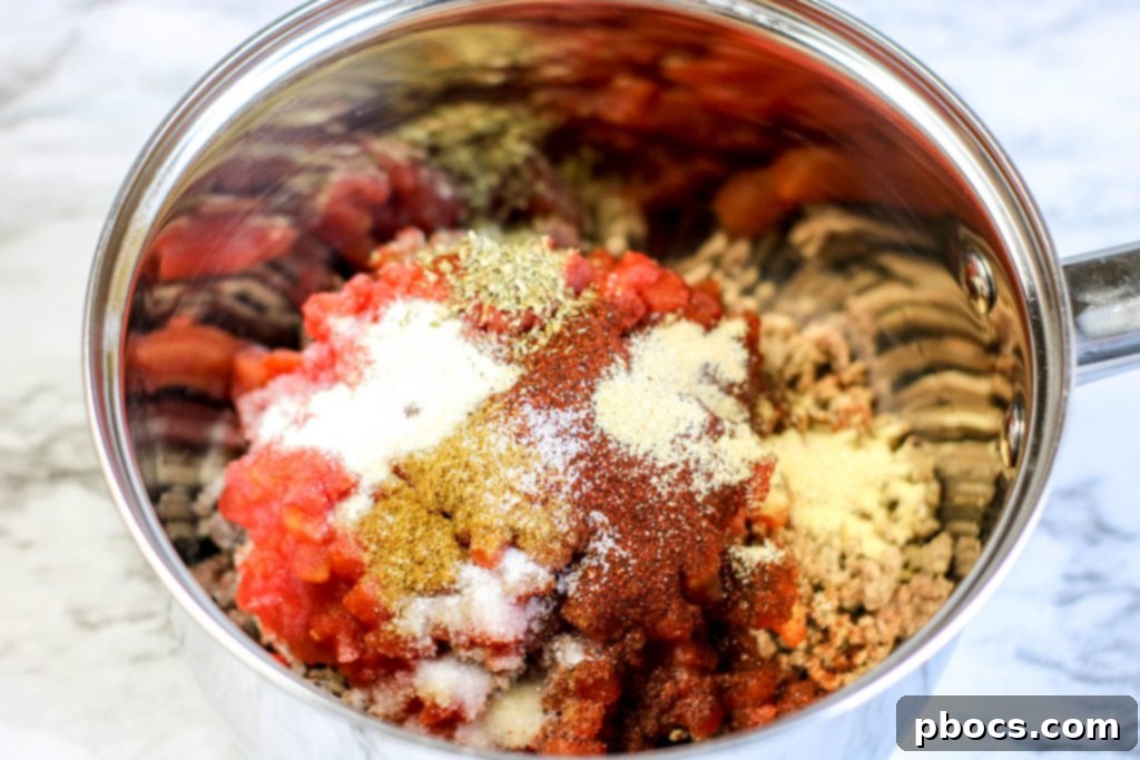 Cooked ground beef in a saucepan with spices and diced tomatoes for chili.