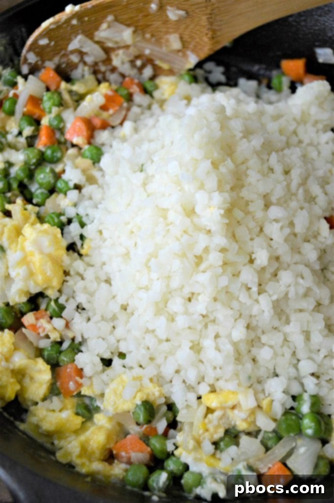 Scrambled eggs being added to the side of sautéed vegetables in a skillet