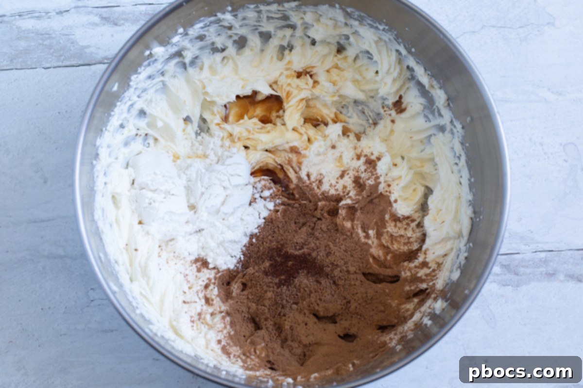 Ingredients being added to the mixing bowl with beaten cream cheese and butter, including cocoa powder and spices.