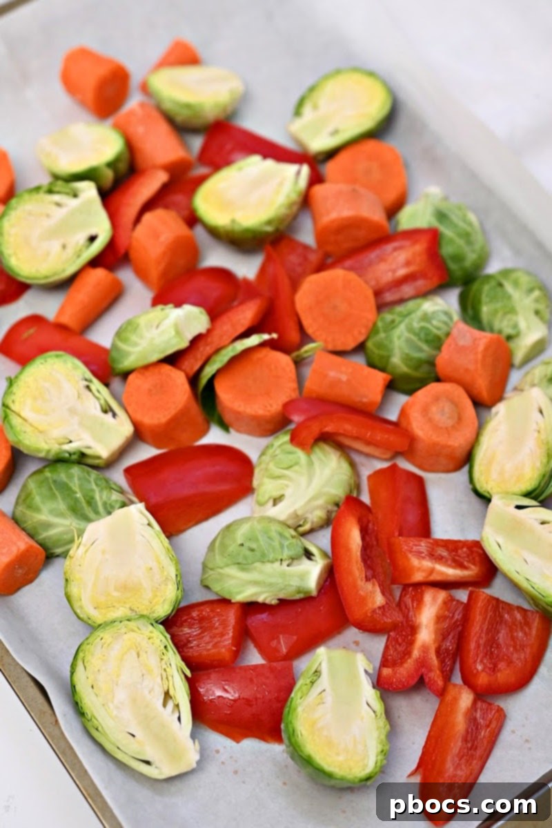 Chopped red bell peppers, carrots, and Brussels sprouts neatly arranged on a baking sheet, drizzled with olive oil and seasoned, ready for the oven.