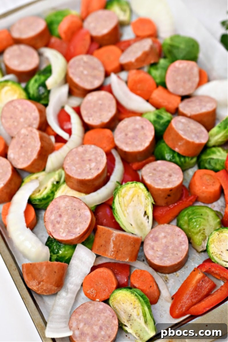 Uncooked slices of natural smoked sausage and onion pieces are added to the sheet pan with partially roasted vegetables, ready for the next stage of cooking.